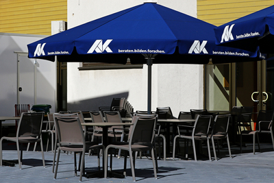 Bild vom Außenbereich vor dem Eingang des Bildungszentrum mit Tischen und Stühlen und einem blauen Sonnenschirm, auf dem Arbeitskammer steht